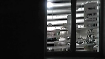 Peeping. Voyeur. Neighbor pervert voyeur in evening on street looks out window as naked sexy neighbor in kitchen prepares dinner for her husband. Naked in public. Naked at home. Family.  Outdoor